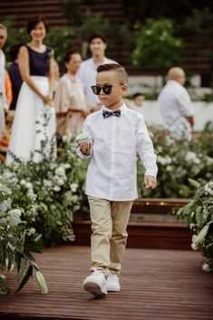 a young boy wearing sunglasses and a bow tie walks down a wooden walkway with people in the background