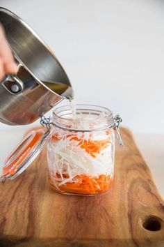 someone is adding shredded carrots into a mason jar on a wooden cutting board with a metal strainer