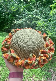 a hand holding up a crocheted hat in front of green grass and trees