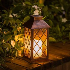 a lit lantern sitting on top of a wooden table in front of some bushes and flowers