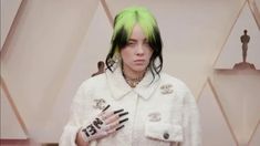 a woman with green hair wearing a white coat and gloves in front of an oscars red carpet