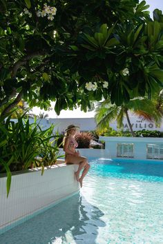 a woman sitting on the edge of a swimming pool next to a lush green tree