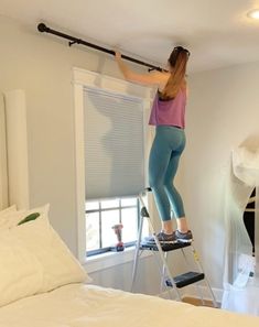 a woman standing on a ladder in front of a window while painting the wall with rollers