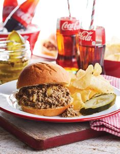 a plate with a sandwich and some chips on the table next to cokes, pickles and soda glasses