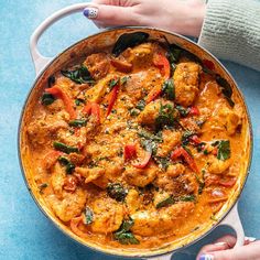 two hands holding a pan filled with chicken and spinach curry on top of a blue table