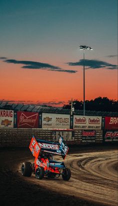 two dirt racing cars going around a track at dusk with the sun setting in the background