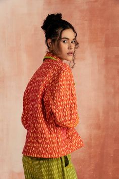 a woman standing in front of a pink wall wearing an orange shirt and green pants