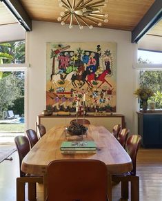 a dining room table with chairs and a painting on the wall behind it in front of windows