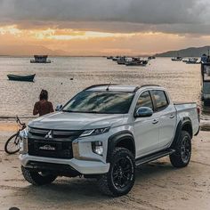 a silver truck parked on top of a sandy beach next to the ocean with boats in the background