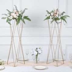 three tall vases with flowers and greenery on top of a table in front of a white wall