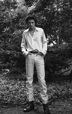 black and white photograph of a man standing in front of trees wearing a shirt and pants