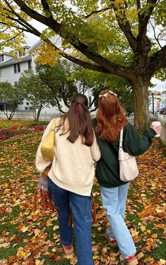 two women walking in the park with their backs to each other as they walk through leaves