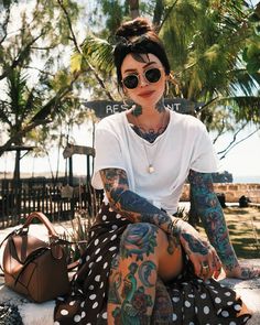 a woman with tattoos sitting on the ground next to a bag and purse in her hand