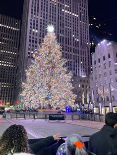 people looking at a large christmas tree in the middle of a city with tall buildings