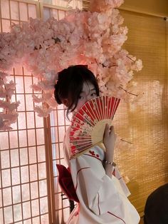 a woman dressed in traditional japanese garb holding a red and white fan next to pink flowers