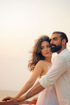 a man and woman standing next to each other near the ocean with their arms around each other