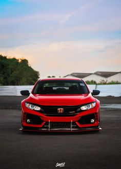 the front end of a red honda civic type car on an asphalt road with trees in the background