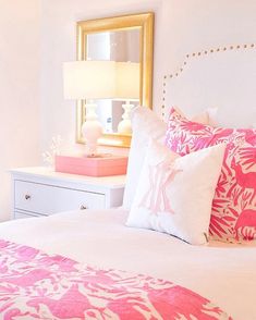 a white bed topped with pink and white pillows next to a dresser under a mirror