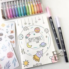 an open notebook next to markers and pens on a white surface with space themed stickers