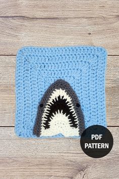 a crocheted blue and white square with a shark in the middle, on top of a wooden surface