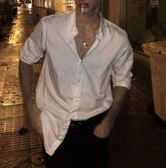 a man in white shirt and black pants standing on the street at night with his hands in his pockets