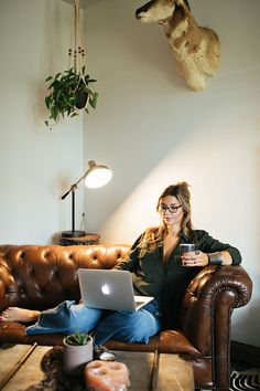 a woman sitting on a couch with a laptop in front of her and a deer head hanging from the wall