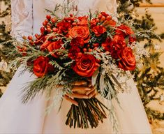 a bride holding a bouquet of red roses and greenery on her wedding day in front of a christmas tree