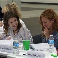 two women are sitting at a table with water bottles and papers on it, smiling