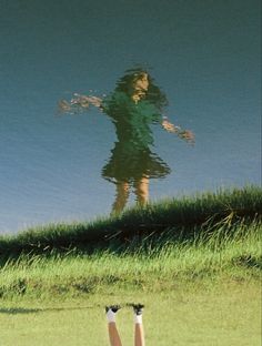 a woman standing on top of a grass covered field next to a lake with her reflection in the water