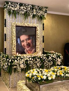 a man standing in front of a photo frame with flowers on the ground and people looking at it