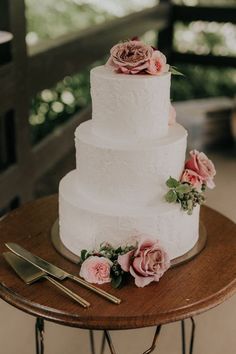 a white wedding cake with pink flowers and greenery sits on a small wooden table