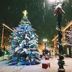 a christmas tree in the middle of a snowy street with lights on and snow falling all around