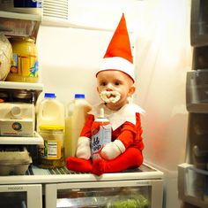 a baby in an elf costume sitting on top of a refrigerator with milk and yogurt