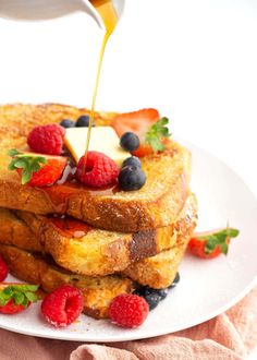 french toast with fruit and syrup being poured onto it on a white plate next to pink napkins