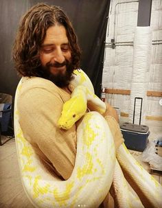 a man with long hair holding a large snake in his arms and smiling at the camera