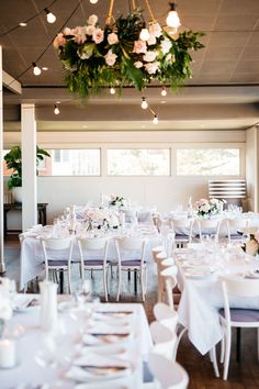 a room filled with lots of tables covered in white tablecloths and flowers hanging from the ceiling