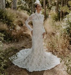 a woman standing in the woods wearing a long sleeved wedding dress with an open back