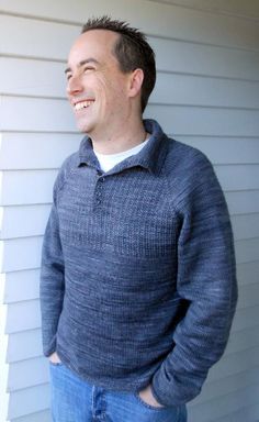 a man standing in front of a house wearing a blue sweater and jeans with his hands in his pockets