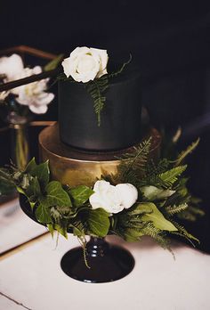 a black and gold wedding cake with white flowers on the top is surrounded by greenery