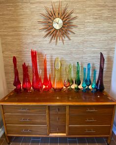 a wooden table topped with lots of colorful glass vases next to a wall clock