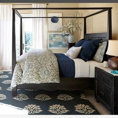 a bedroom with a four poster bed, blue and white comforter, black nightstands, and large windows