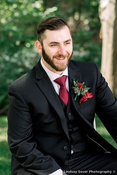 a man in a suit and tie sitting on a bench smiling at the camera while wearing a red rose boutonniere