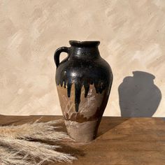 a black and brown vase sitting on top of a wooden table next to dry grass