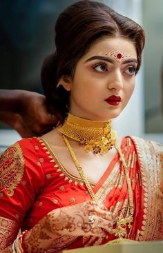 a woman in a red and gold sari with jewelry on her neck, looking at the camera