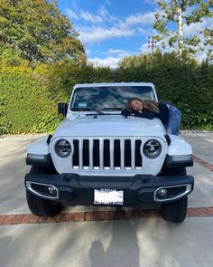 a woman leaning on the hood of a white jeep parked in front of a hedge