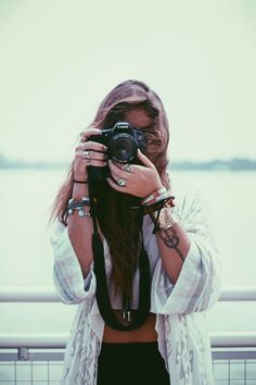 a woman holding a camera up to her face while standing next to a body of water