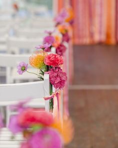 colorful flowers are lined up along the back of white chairs at a wedding ceremony in an orange and pink color scheme