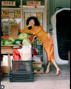 a woman in an orange dress leaning on a table with watermelons and other produce