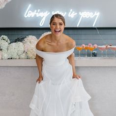 a woman in a white dress standing next to a wall with flowers and drinks on it