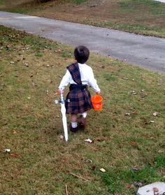 a little boy in a kilt walking down the street with an orange cup and white baseball bat